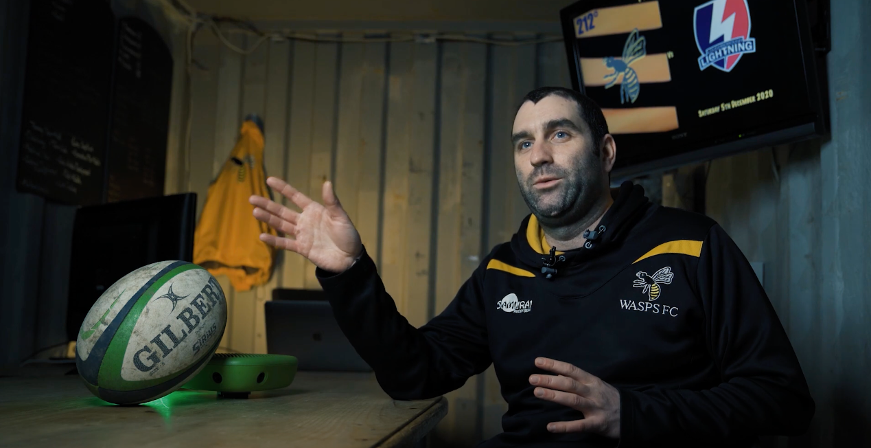A thumbnail for the article 'How Veo Became a Game-Changer for Wasps FC Women's Team', featuring a coach sitting next to a table with a rugby ball and a television screen displaying the Wasps FC logo in the background.