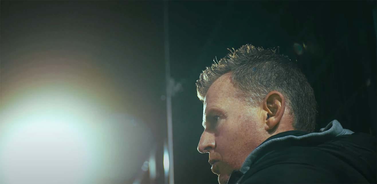 A photo of rugby icon Nigel Owens, shot from the left side with a dark background behind him.