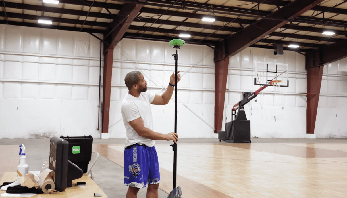 Photo of a basketball player setting up a tripod with a Veo camera on a basketball court, with one basketball basket visible in the background, preparing to record a game or training session for advanced video analysis.