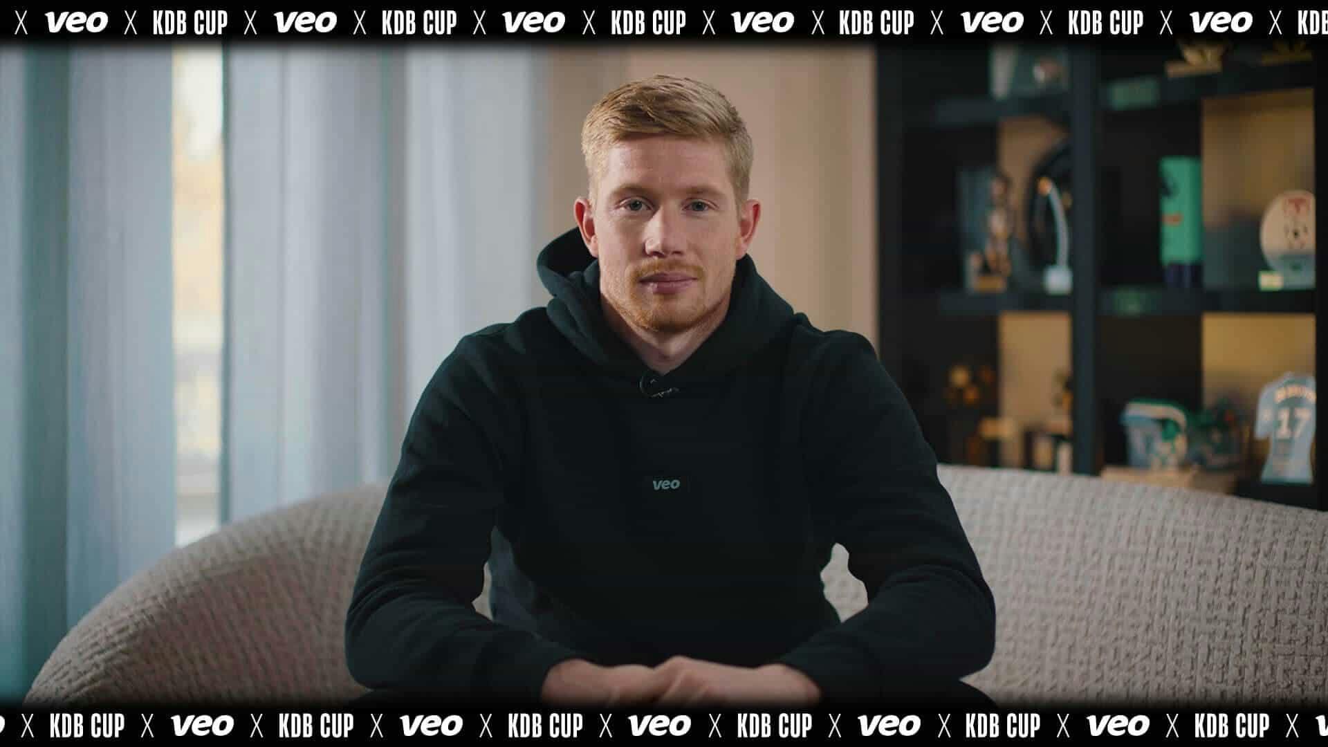 Photo of Kevin de Bruyne sitting comfortably on a couch while holding a Veo soccer camera in his hand. He is dressed in casual sportswear and has a focused expression on his face as he looks at the camera