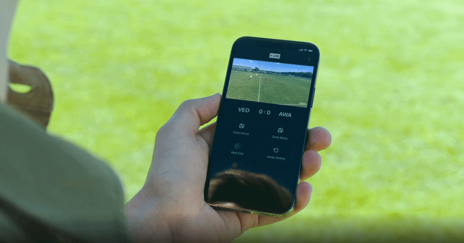 A person holding a mobile phone and interacting with the Veo Live app on the football field. The app allows users to easily set up goals, halftime, and end of game markers during live games